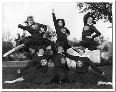 A black and white photo of a group of cheerleaders. 3 women are standing in the foreground while 3 other women are mid jump.