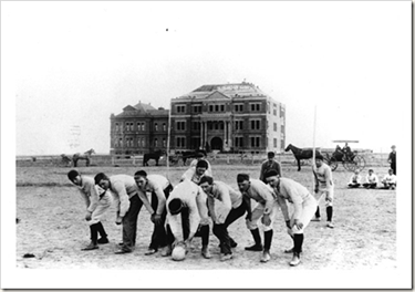 A black and white photo of a group of young men standing at the line of scrimmage on a football field. In the background a large building can be seen along with a few horses.